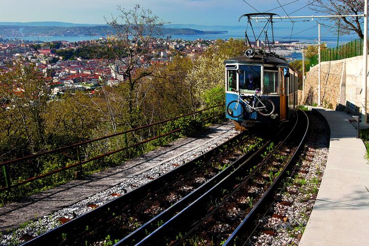 Eine blaue Straßenbahn, die auf einen Hügel fährt, mit der Stadt Triest im Hintergrund
