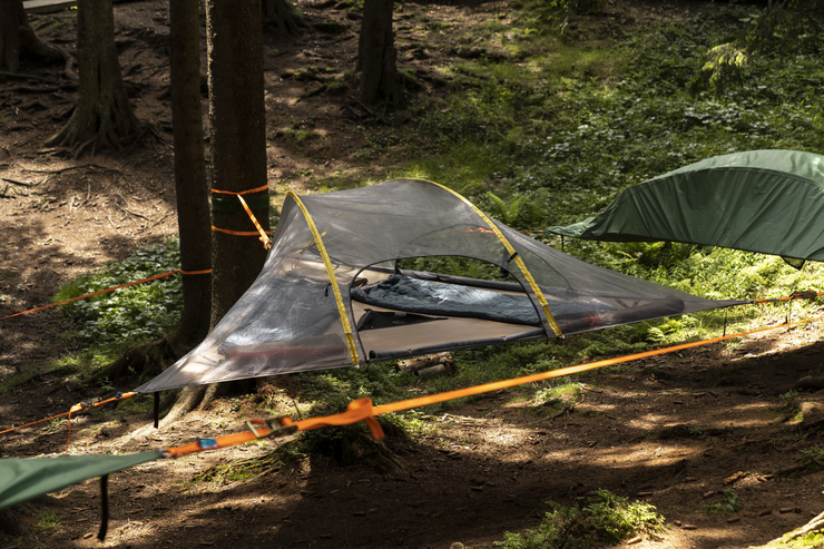Ein schwebendes Zelt, zwischen Bäumen in einem Wald aufgehängt, auf einem Netz gespannt.