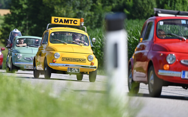 Kleine bunte Autos fahren auf einer Landstraße. Eines der Autos ist gelb und hat ein ÖAMTC-Schild auf dem Dach.