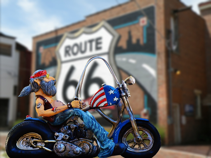Am Foto ist ein riesiges Schild der legendären US-Straße Route 66 zu sehen, mit einer bunten Figur im Vordergrund, die eine Harley Davidson und einen Biker in Jeans und rotem Stirntuch zeigt.