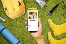 Eine Hand hält ein Smartphone mit einer "Festival"-Checkliste. Um das Smartphone herum liegen ein Rucksack, eine Kamera, ein Mikrofon, gelbe Gummistiefel, eine blaue Isomatte und ein Festival-Armband.