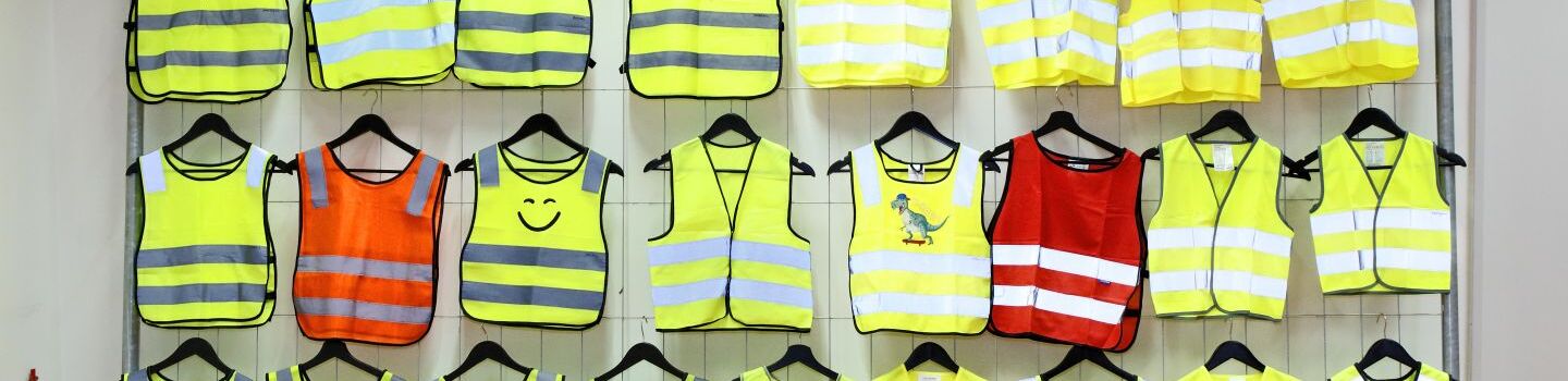 25 gelbe und orange Warnwesten hängen an einem Zaun. Die Reflektoren sind bei manchen erkennbar.