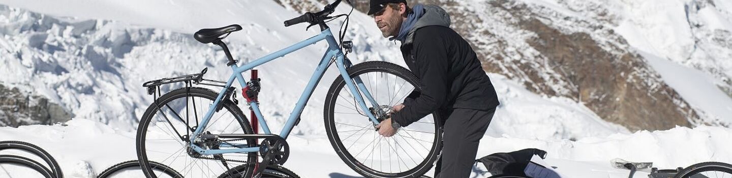 Ein Mann steht im Gebirge im Schnee und repariert ein blaues Fahrrad auf einem Fahrradständer. Er ist von mehreren Fahrradreifen umgeben und im Hintergrund sind schneebedeckte Berge zu sehen.