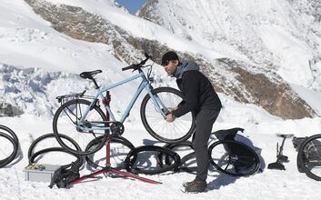 Ein Mann steht im Gebirge im Schnee und repariert ein blaues Fahrrad auf einem Fahrradständer. Er ist von mehreren Fahrradreifen umgeben und im Hintergrund sind schneebedeckte Berge zu sehen.