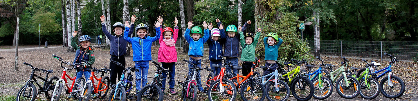14 Kinderfahrräder sind auf einem Waldweg aufgestellt, dahinter einige Kinder mit Radhelm, die die Hände in die Höhe heben.