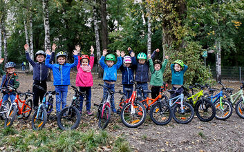 14 Kinderfahrräder sind auf einem Waldweg aufgestellt, dahinter einige Kinder mit Radhelm, die die Hände in die Höhe heben.