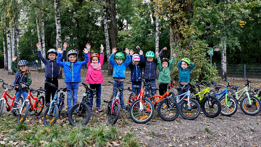 14 Kinderfahrräder sind auf einem Waldweg aufgestellt, dahinter einige Kinder mit Radhelm, die die Hände in die Höhe heben.