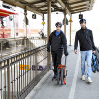 Das Bild zeigt zwei Männer mit zusammengeklappten Fahrrädern auf einem Bahnsteig. Vom Betrachter aus gesehen befindet sich rechts der Männer das Bahngleis, links ein Geländer. Der vom Betrachter aus gesehen linke Mann ist mit einer dunklen Hose und einer dunklen Jacke bekleidet und hat ein oranges Klapprad. Der vom Betrachter aus gesehen rechte Mann ist mit einer hellblauen Hose und einer schwarzen Jacke bekleidet und hat ein blaues Klapprad. Beide Männer tragen Radhelme.