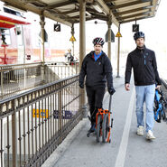 Das Bild zeigt zwei Männer mit zusammengeklappten Fahrrädern auf einem Bahnsteig. Vom Betrachter aus gesehen befindet sich rechts der Männer das Bahngleis, links ein Geländer. Der vom Betrachter aus gesehen linke Mann ist mit einer dunklen Hose und einer dunklen Jacke bekleidet und hat ein oranges Klapprad. Der vom Betrachter aus gesehen rechte Mann ist mit einer hellblauen Hose und einer schwarzen Jacke bekleidet und hat ein blaues Klapprad. Beide Männer tragen Radhelme.