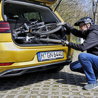 Das Bild zeigt einen Mann, der ein zusammengeklapptes Fahrrad in den Kofferraum eines Autos verlädt. Der Mann hockt vor dem geöffneten Kofferraum. Er trägt eine blaue Hose, eine dunkle Jacke und einen Radhelm. Das Auto ist dunkelgelb und hat ein Kennzeichen der Stadt München in Deutschland.