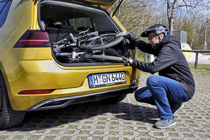 Das Bild zeigt einen Mann, der ein zusammengeklapptes Fahrrad in den Kofferraum eines Autos verlädt. Der Mann hockt vor dem geöffneten Kofferraum. Er trägt eine blaue Hose, eine dunkle Jacke und einen Radhelm. Das Auto ist dunkelgelb und hat ein Kennzeichen der Stadt München in Deutschland.