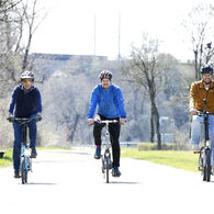 Das Bild zeigt drei Männer auf Fahrrädern. Sie fahren bei Sonnenschein nebeneinander eine Straße entlang. Alle drei Männer tragen Radhelme. Der vom Betrachter des Bildes aus gesehen linke der drei sitzt auf einem hellblauen Fahrrad und ist mit einer dunkelblauen Hose und einer dunkelblauen Jacke bekleidet. Der mittlere der drei Männer sitzt auf einem silbernen Fahrrad und ist mit einer schwarzen Hose sowie einer blauen Jacke bekleidet. Der vom Betrachter aus gesehen rechte der drei Männer sitzt auf einem schwarzen Fahrrad und ist mit einer schwarzen Hose und einer hellbraunen Jacke bekleidet.