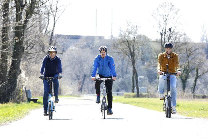 Das Bild zeigt drei Männer auf Fahrrädern. Sie fahren bei Sonnenschein nebeneinander eine Straße entlang. Alle drei Männer tragen Radhelme. Der vom Betrachter des Bildes aus gesehen linke der drei sitzt auf einem hellblauen Fahrrad und ist mit einer dunkelblauen Hose und einer dunkelblauen Jacke bekleidet. Der mittlere der drei Männer sitzt auf einem silbernen Fahrrad und ist mit einer schwarzen Hose sowie einer blauen Jacke bekleidet. Der vom Betrachter aus gesehen rechte der drei Männer sitzt auf einem schwarzen Fahrrad und ist mit einer schwarzen Hose und einer hellbraunen Jacke bekleidet.