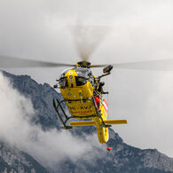 ÖAMTC-Flugrettung_C14_Pilotprojekt mit Rettungswinde Das Bild zeigt einen gelben Hubschrauber im Flug, im Hintergrund felsiges Gebirge. Der Hubschrauber ist mit einer Seilwinde ausgestattet, ein Besatzungsmitglied steht außen an der Maschine und hält das Seil.