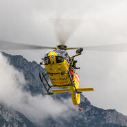 Das Bild zeigt einen gelben Hubschrauber im Flug, im Hintergrund felsiges Gebirge. Der Hubschrauber ist mit einer Seilwinde ausgestattet, ein Besatzungsmitglied steht außen an der Maschine und hält das Seil.