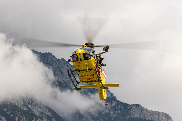 Das Bild zeigt einen gelben Hubschrauber im Flug, im Hintergrund felsiges Gebirge. Der Hubschrauber ist mit einer Seilwinde ausgestattet, ein Besatzungsmitglied steht außen an der Maschine und hält das Seil.