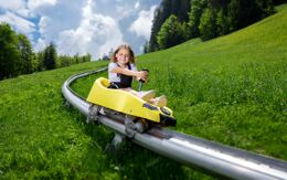 Kind sitzt in einer gelben Sommerrodelbahn und fährt lächelnd über Schienen auf einer Bergwiese.