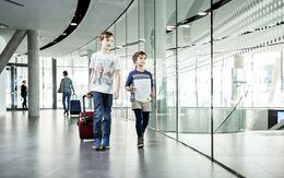 ein kleines Mädchen und ein kleiner Junge gehen einen Gang entlang. Der Junge zieht einen Trolley, das Mädchen hat ein Dokument in der Hand.