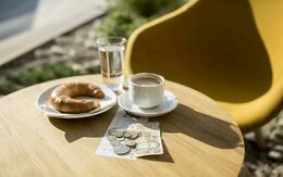 Ein kleiner runder Tisch im freien. Auf dem Tisch steht eine weiße Tasse mit Kaffee, ein Glas Wasser und ein Teller mit einem Croissant. Auf dem Tisch liegen auch mehrere Münzen und ein Geldschein. Im Hintergrund ist ein gelber Stuhl zu sehen.