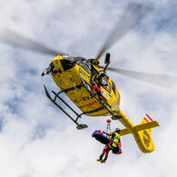 ÖAMTC-Flugrettung_C14_Pilotprojekt mit Rettungswinde Das Bild zeigt einen gelben Hubschrauber im Flug. Der Hubschrauber ist mit einer Seilwinde ausgestattet, ein Besatzungsmitglied steht außen an der Maschine, ein weiteres Besatzungsmitglied wird gerade vom Hubschrauber abgeseilt.