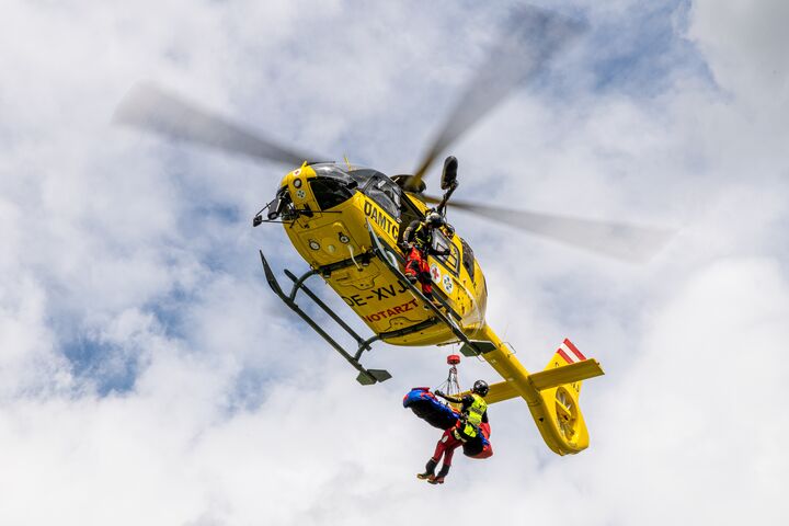Das Bild zeigt einen gelben Hubschrauber im Flug. Der Hubschrauber ist mit einer Seilwinde ausgestattet, ein Besatzungsmitglied steht außen an der Maschine, ein weiteres Besatzungsmitglied wird gerade vom Hubschrauber abgeseilt.