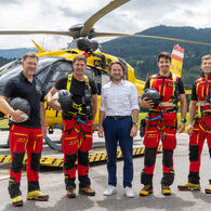 ÖAMTC-Flugrettung_C14_Pilotprojekt mit Rettungswinde Auf dem Bild sieht man fünf Männer vor einem gelben Hubschrauber. Im Hintergrund eine grüne Wiese und eine hügelige, bewaldete Landschaft. Die beiden jeweils links und rechts außen stehenden Männer gehören zur Einsatzcrew und tragen die rot-schwarze Uniform der ÖAMTC Flugrettung. Der Mann in der Mitte ist der Geschäftsführer der ÖAMTC Flugrettung und trägt eine blaue Hose und ein weißes Hemd.