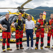 Auf dem Bild sieht man fünf Männer vor einem gelben Hubschrauber. Im Hintergrund eine grüne Wiese und eine hügelige, bewaldete Landschaft. Die beiden jeweils links und rechts außen stehenden Männer gehören zur Einsatzcrew und tragen die rot-schwarze Uniform der ÖAMTC Flugrettung. Der Mann in der Mitte ist der Geschäftsführer der ÖAMTC Flugrettung und trägt eine blaue Hose und ein weißes Hemd.