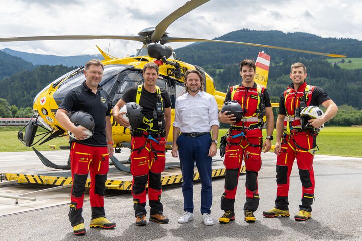 Auf dem Bild sieht man fünf Männer vor einem gelben Hubschrauber. Im Hintergrund eine grüne Wiese und eine hügelige, bewaldete Landschaft. Die beiden jeweils links und rechts außen stehenden Männer gehören zur Einsatzcrew und tragen die rot-schwarze Uniform der ÖAMTC Flugrettung. Der Mann in der Mitte ist der Geschäftsführer der ÖAMTC Flugrettung und trägt eine blaue Hose und ein weißes Hemd.