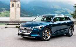 Ein blauer Audi e-Tron, Ansicht seitlich von vorne, steht auf einer Straße, im Hintergrund sind ein Turm, Wasser und Berge.