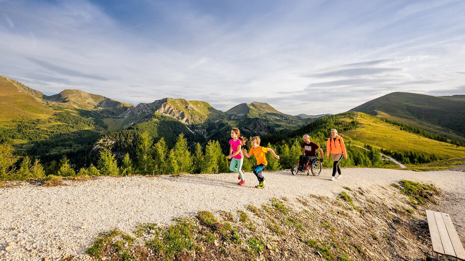 Wanderweg in den Bergen in Kärnten. Zwei Kinder laufen vor, die Erwachsenen, ein Mann im Rollstuhl und eine Frau folgen ihnen.