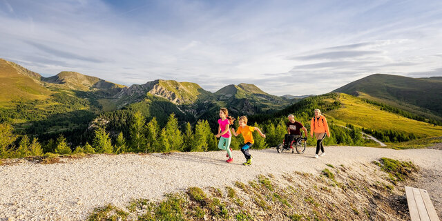 Wanderweg in den Bergen in Kärnten. Zwei Kinder laufen vor, die Erwachsenen, ein Mann im Rollstuhl und eine Frau folgen ihnen.