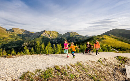 Wanderweg in den Bergen in Kärnten. Zwei Kinder laufen vor, die Erwachsenen, ein Mann im Rollstuhl und eine Frau folgen ihnen.