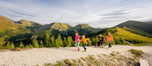 Wanderweg in den Bergen in Kärnten. Zwei Kinder laufen vor, die Erwachsenen, ein Mann im Rollstuhl und eine Frau folgen ihnen.