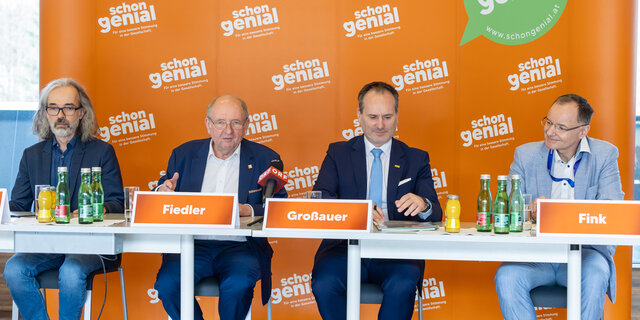 Vier Herren im Anzug sitzen an einem Tisch und geben eine Pressekonferenz