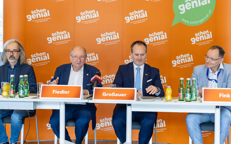 Vier Herren im Anzug sitzen an einem Tisch und geben eine Pressekonferenz