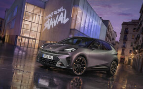 Ein grauer Cupra Raval steht vor einem modernen Gebäude, auf dem ein "Raval"-Schriftzug leuchtet.