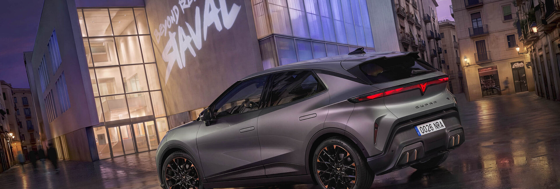 Ein grauer Cupra Raval steht vor einem modernen Gebäude, auf dem ein "Raval"-Schriftzug leuchtet.