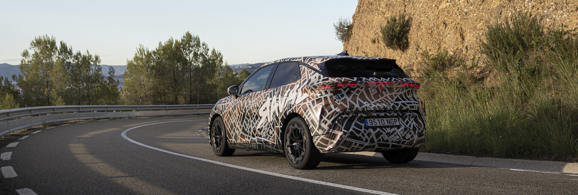Ein mit Tarnfolie folierter Cupra Raval fährt um eine Kurve auf einer Landstraße.
