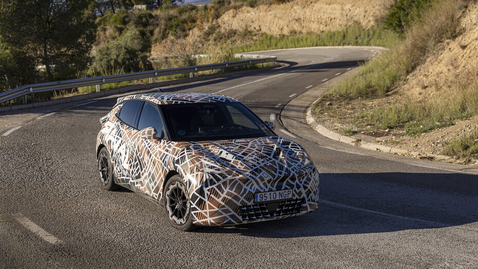 Ein mit Tarnfolie folierter Cupra Raval fährt um eine Kurve auf einer Landstraße.