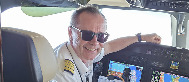 Flugkapitän Harry Gruber sitzt im Cockpit eines Flugzeugs und lächelt. Er hat kurze graue Haare und eine schwarze Sonnenbrille. Er trägt eine Pilotenuniform.