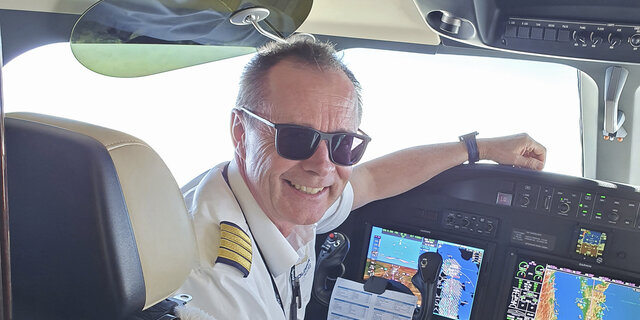 Flugkapitän Harry Gruber sitzt im Cockpit eines Flugzeugs und lächelt. Er hat kurze graue Haare und eine schwarze Sonnenbrille. Er trägt eine Pilotenuniform.