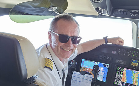 Flugkapitän Harry Gruber sitzt im Cockpit eines Flugzeugs und lächelt. Er hat kurze graue Haare und eine schwarze Sonnenbrille. Er trägt eine Pilotenuniform.
