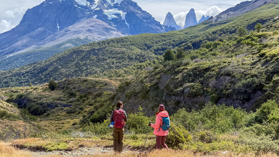 zwei Wanderer blicken von einer Lichtung auf die drei Granit-Berge Tres Torres del Paine, die hinter iem Hügel hervorschauen.