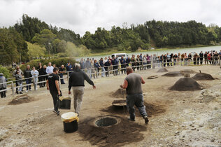 Männer holen aus einem heißen Erdloch einen Topf. Es schauen viele Touristen zu.