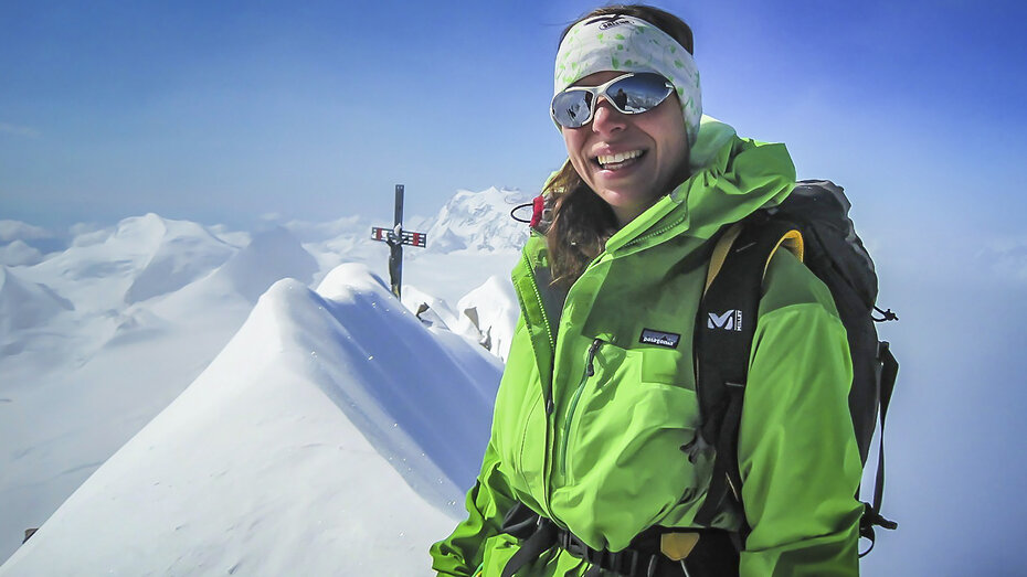 Eine Frau im grünen Wintergewand steht am Berg, im Hintergrund ein Gipfelkreuz. 