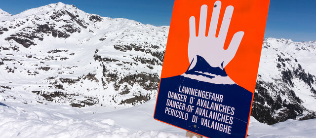 Ein rotes Lawinengefahr-Schild, man sieht eine weiße Hand, die "Halt" signalisiert. Dahinter sind die Berge zu sehen.