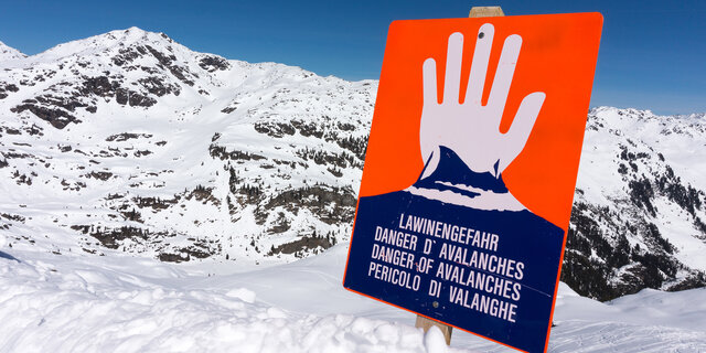 Ein rotes Lawinengefahr-Schild, man sieht eine weiße Hand, die "Halt" signalisiert. Dahinter sind die Berge zu sehen.