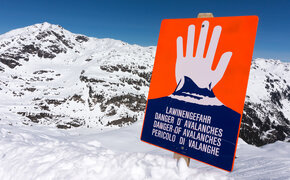 Ein rotes Lawinengefahr-Schild, man sieht eine weiße Hand, die "Halt" signalisiert. Dahinter sind die Berge zu sehen.