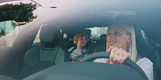 Eine blonde Frau am Steuer eines Autos, am Rücksitz sitzt ein Kind.