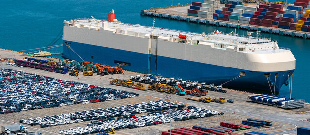 Tausende Autos auf einem Parkplatz an einem Hafen, ein Transportschiff liegt vor Anker.
